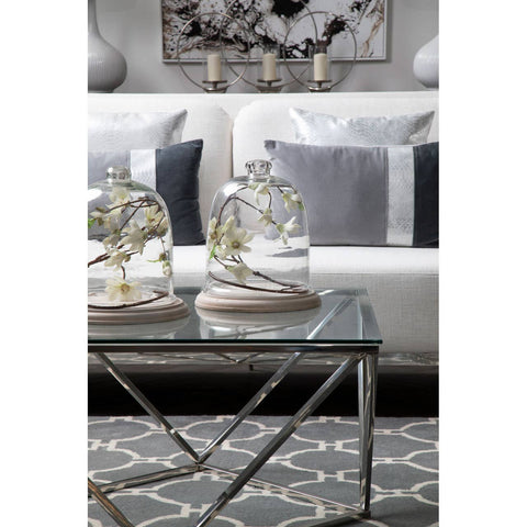 A stylish two-seater sofa in ivory and mink upholstery, positioned in a modern living room setting. The sofa features plush cushions and is complemented by a glass coffee table with decorative items, including two glass cloches and a patterned area rug beneath. The background showcases a piece of wall art and soft lighting, creating an inviting atmosphere
