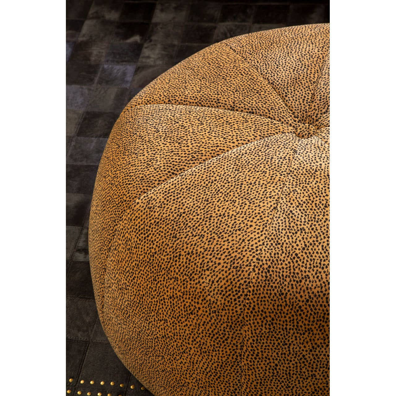 Round pouffe featuring a textured leopard print design in shades of brown and tan, with a tufted top and a soft, plush surface. The background is a dark, woven material that contrasts with the pouffe's color