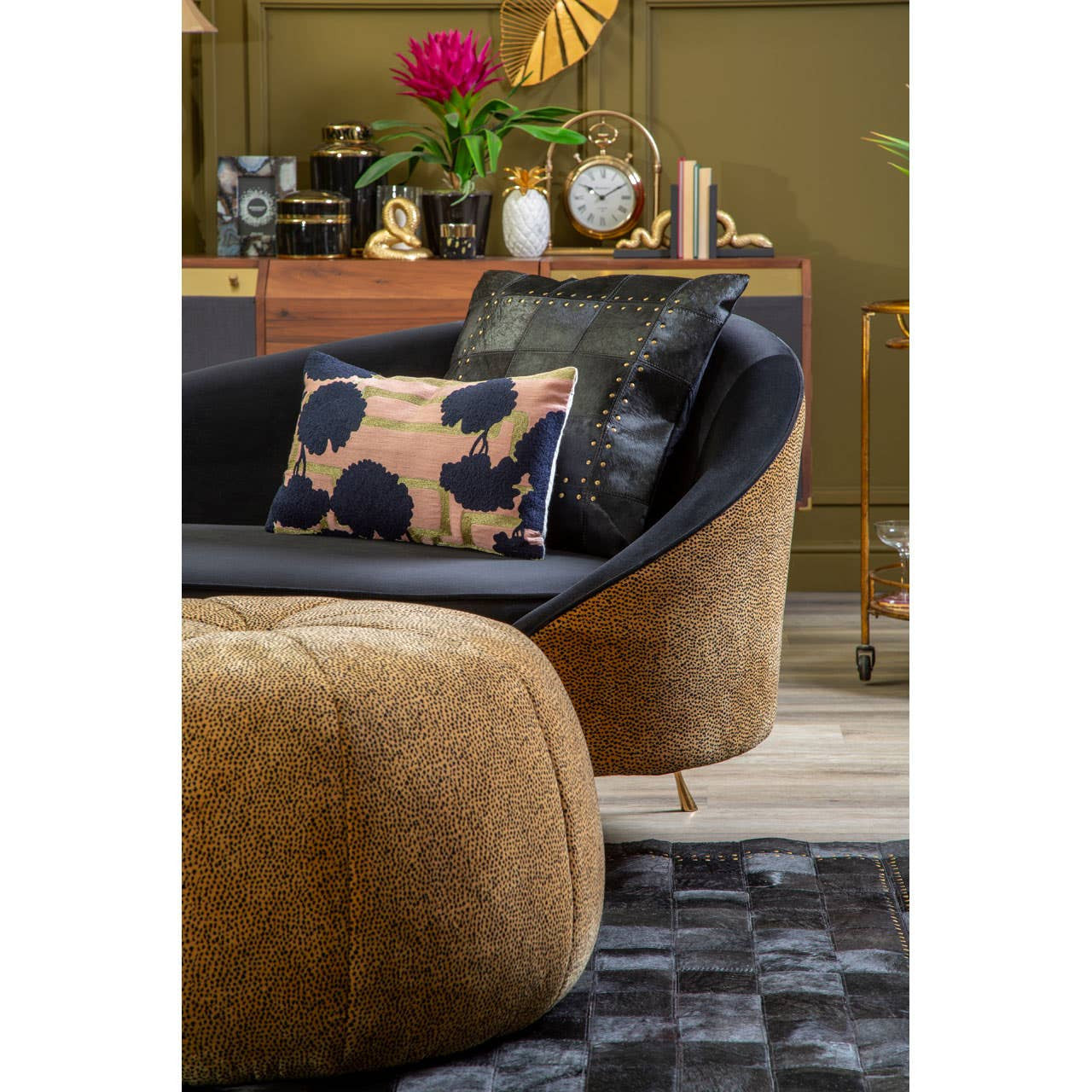 A leopard print pouffe in a warm brown color sits in a stylish living room setting, accompanied by a black and gold accent chair and a decorative pillow featuring a floral design. The background includes a wooden console table adorned with decorative items and a vibrant pink flower arrangement. The floor is covered with a textured rug, adding depth to the cozy atmosphere