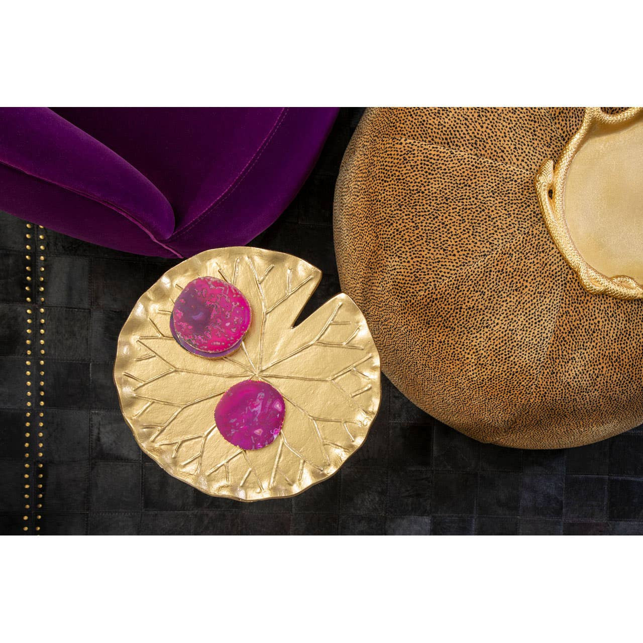 A leopard print pouffe in a warm beige color, featuring a textured surface, is positioned next to a round, gold decorative table with two pink coasters on top. The background includes a dark surface, and a plush purple cushion is partially visible