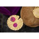 A leopard print pouffe in a warm beige color, featuring a textured surface, is positioned next to a round, gold decorative table with two pink coasters on top. The background includes a dark surface, and a plush purple cushion is partially visible