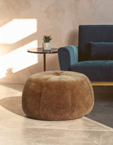 Round pouffe with a leopard print design in shades of brown, placed on a light-colored floor. A blue sofa and a small side table with a plant are visible in the background