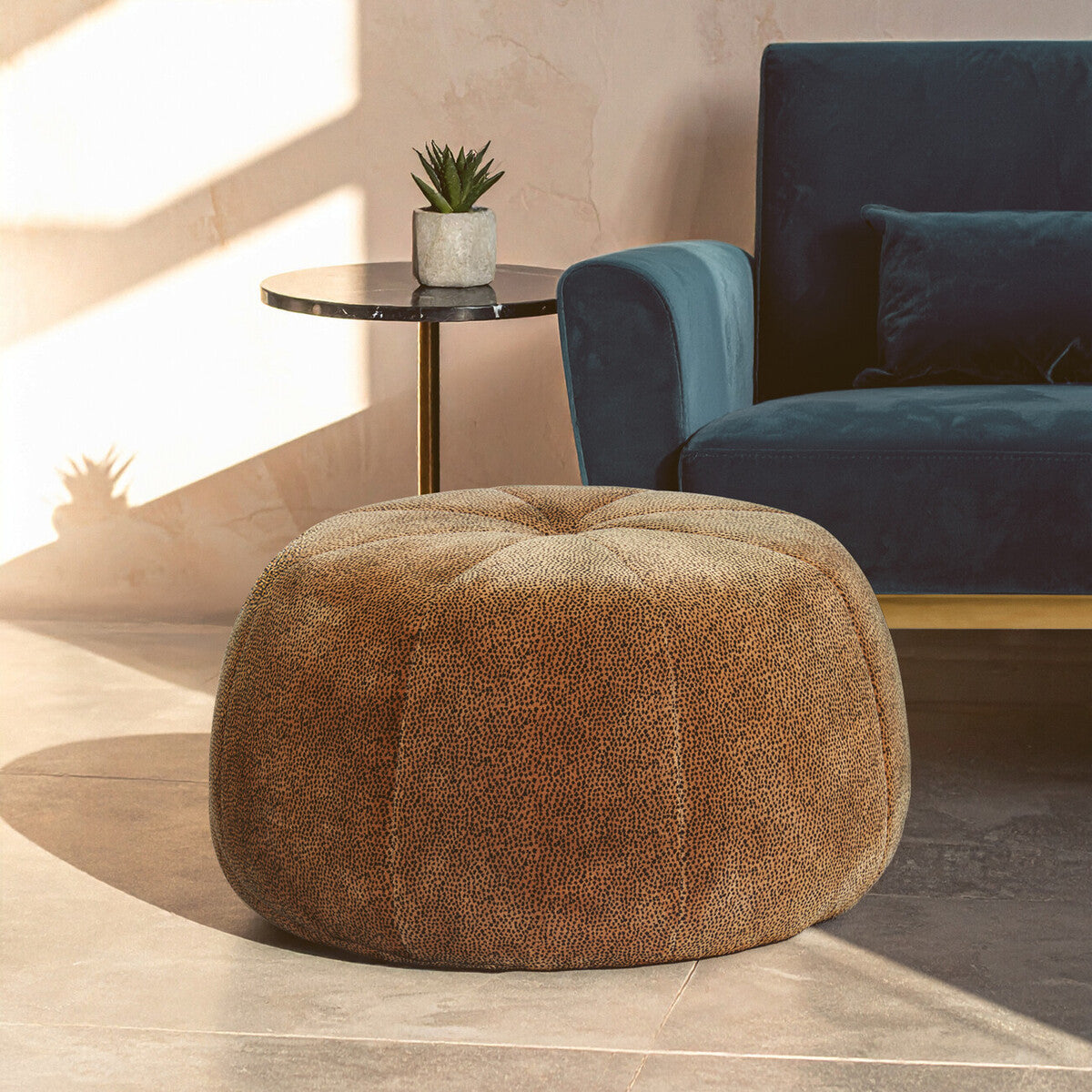 Round pouffe with a soft, leopard print fabric in shades of brown, placed on a light-colored floor. A small potted plant sits on a side table nearby, with a blue upholstered sofa in the background