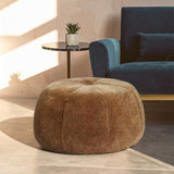 Round pouffe with a soft, leopard print fabric in shades of brown, placed on a light-colored floor. A small potted plant sits on a side table nearby, with a blue upholstered sofa in the background
