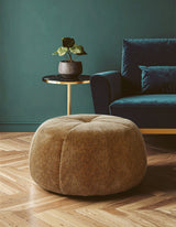 Round pouffe with a leopard print pattern in a warm brown color, placed on a wooden floor. A small side table with a potted plant is visible next to the pouffe, and a dark blue sofa is in the background against a green wall