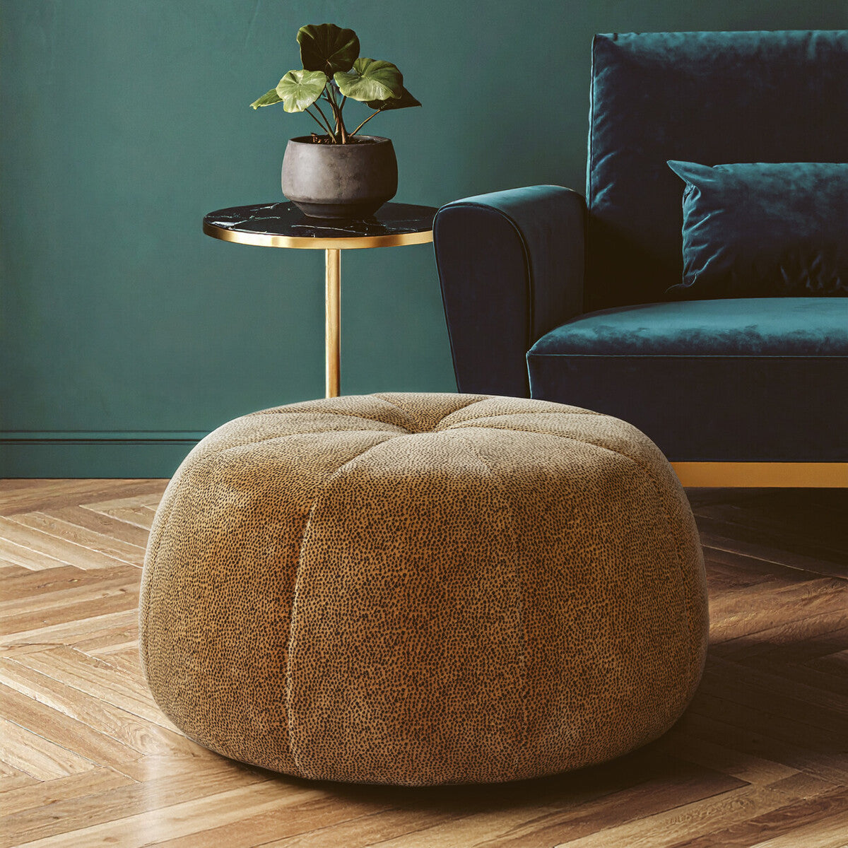 Round pouffe in a leopard print pattern, featuring a soft, textured surface. It is positioned on a wooden floor, next to a small side table with a potted plant. A dark blue sofa is visible in the background against a green wall