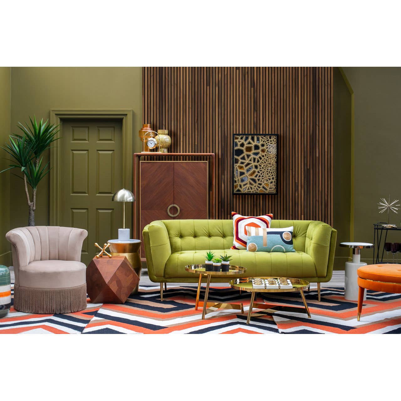 A vibrant orange velvet footstool placed in a stylish living room setting, featuring a green sofa, a patterned rug, and various decorative elements like a lamp and a plant. The footstool complements the colorful decor and adds a cozy touch to the space