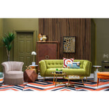 A vibrant orange velvet footstool placed in a stylish living room setting, featuring a green sofa, a patterned rug, and various decorative elements like a lamp and a plant. The footstool complements the colorful decor and adds a cozy touch to the space