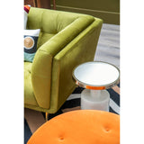 A vibrant orange velvet footstool sits in the foreground, featuring a tufted design. In the background, a green upholstered armchair is partially visible, along with a small round side table topped with a white surface and a metallic rim. The setting includes a patterned rug beneath the furniture