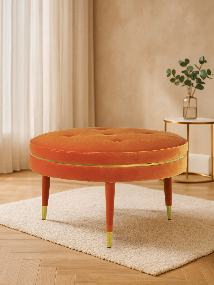 Round orange velvet footstool with tufted top and wooden legs, featuring gold accents at the feet, placed on a light-colored rug in a bright room with natural light and a small potted plant in the background