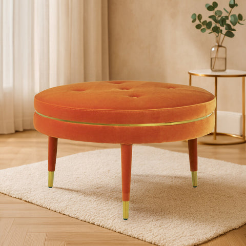 Round orange velvet footstool with a tufted top and wooden legs, featuring gold accents at the feet, placed on a light-colored rug in a bright, modern living space with soft natural light and a potted plant in the background