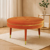 Round orange velvet footstool with a tufted top and wooden legs, featuring gold accents at the feet, placed on a light-colored rug in a bright, modern living space with soft natural light and a potted plant in the background