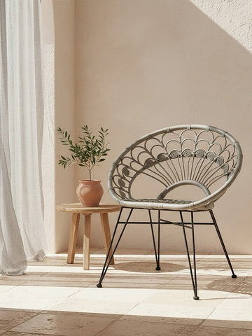 A grey natural rattan chair with a circular design, positioned next to a small wooden side table. A potted plant sits on the table, and soft sunlight filters through sheer curtains, casting gentle shadows on the floor. The background features a warm, neutral wall color