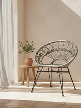 A grey natural rattan chair with a circular design, positioned next to a small wooden side table. A potted plant sits on the table, and soft sunlight filters through sheer curtains, casting gentle shadows on the floor. The background features a warm, neutral wall color