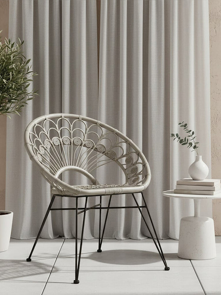 A grey natural rattan chair with a curved backrest and open weave design, supported by black metal legs. The chair is positioned next to a small round side table with a decorative vase and a plant, against a backdrop of light-colored curtains