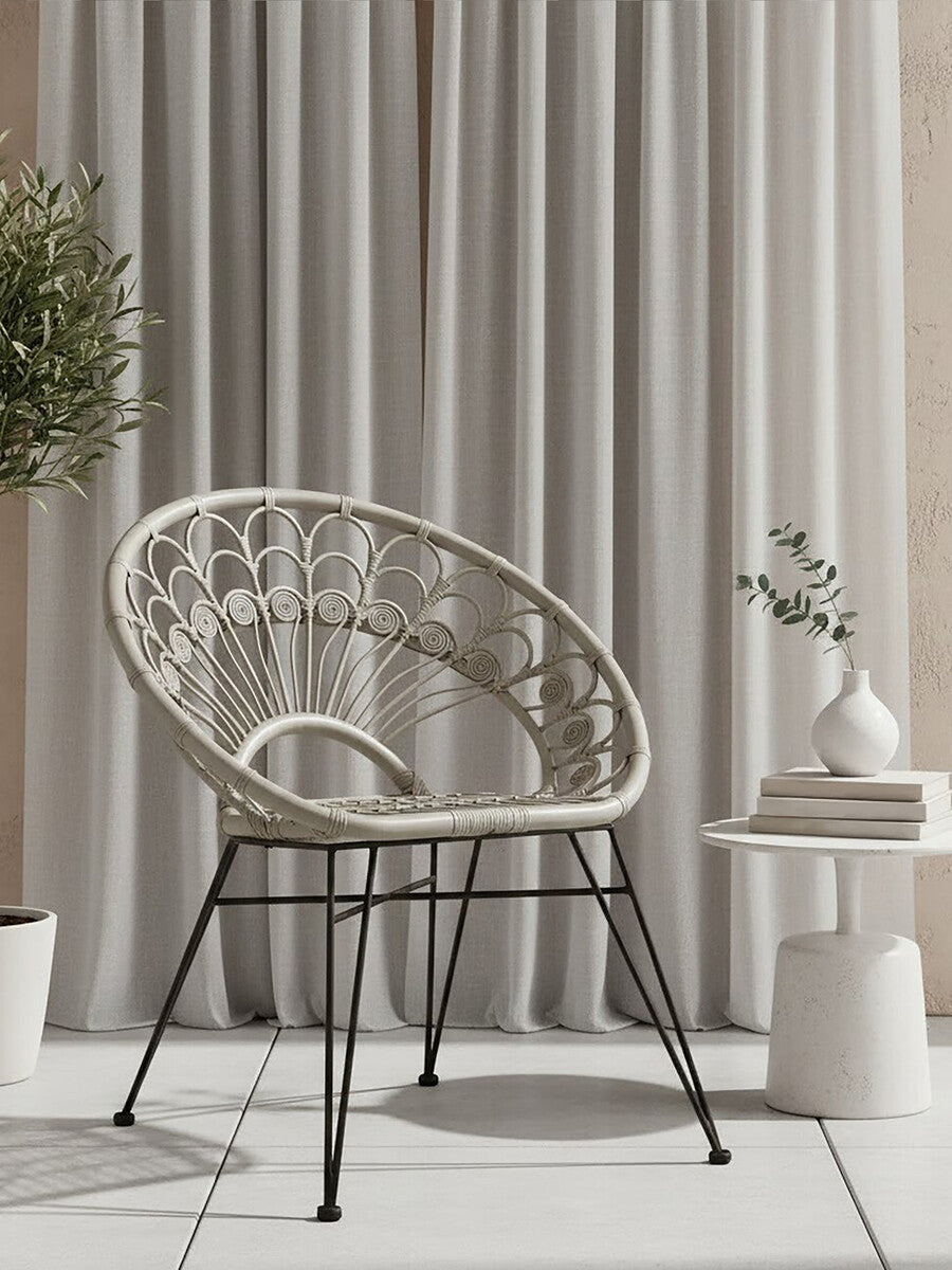 A grey natural rattan chair with a curved backrest and open weave design, supported by black metal legs. The chair is positioned next to a small round side table with a decorative vase and a plant, against a backdrop of light-colored curtains