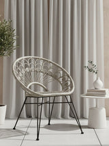 A grey natural rattan chair with a curved backrest and open weave design, supported by black metal legs. The chair is positioned next to a small round side table with a decorative vase and a plant, against a backdrop of light-colored curtains