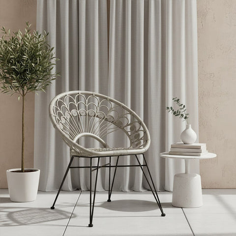 A grey natural rattan chair with a curved design sits next to a small white side table, adorned with a decorative plant and a stack of books. Light grey curtains drape in the background, complementing the neutral tones of the setting