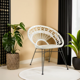 A white natural rattan chair with a circular backrest and black metal legs, positioned in a bright room with beige walls. A large green plant in a textured pot is nearby, and there is a round white rug on the floor. Black curtains frame a window, adding contrast to the light space