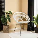 A white natural rattan chair with a curved backrest and metal legs is positioned in a cozy corner, surrounded by a potted plant and a circular rug, against a backdrop of beige walls and black curtains