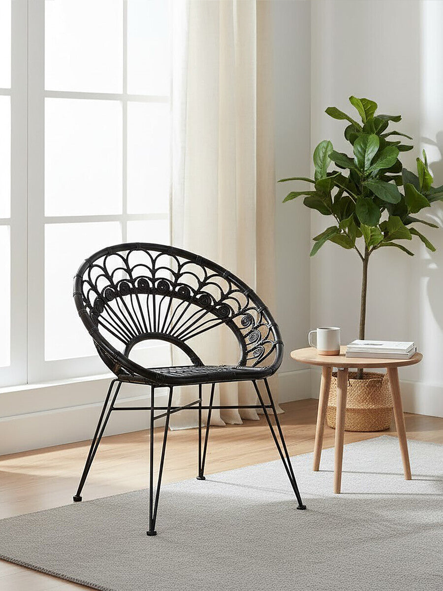 A black natural rattan chair with a circular design and slender legs, positioned next to a small wooden side table in a bright room with large windows and a potted plant in the background