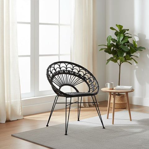 A black natural rattan chair with a circular backrest and slender legs, positioned in a bright room with large windows. A small round wooden side table is next to the chair, and a potted plant adds a touch of greenery to the space. Light-colored curtains frame the windows, enhancing the airy atmosphere
