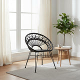 A black natural rattan chair with a circular backrest and slender legs, positioned in a bright room with large windows. A small round wooden side table is next to the chair, and a potted plant adds a touch of greenery to the space. Light-colored curtains frame the windows, enhancing the airy atmosphere