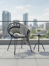 A black natural rattan chair with a circular backrest and four slender legs is positioned on a balcony. Next to the chair, there is a small round table with a potted plant on top. In the background, tall modern buildings and a clear blue sky are visible, creating a bright and airy outdoor setting