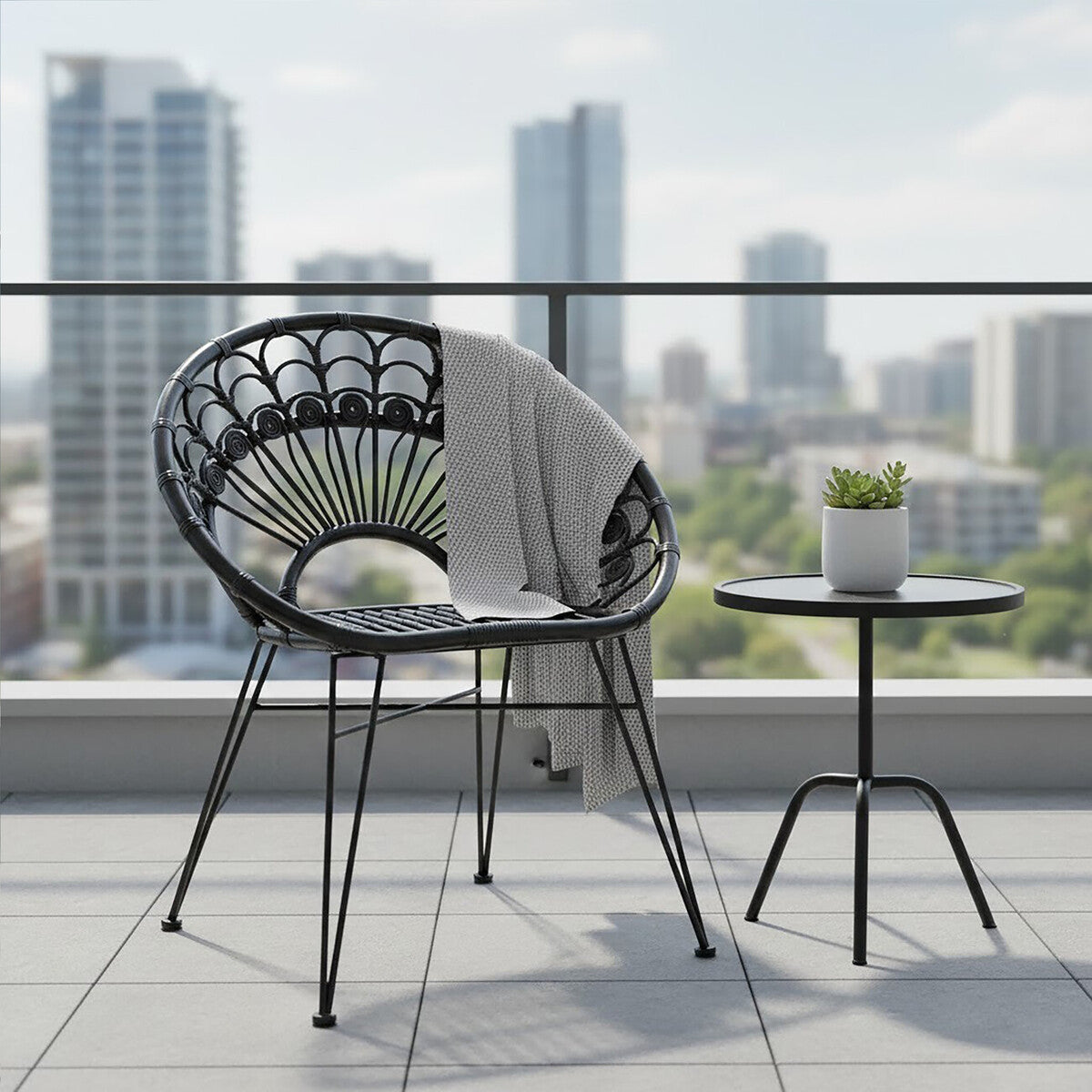 A black natural rattan chair with a curved backrest, placed on a balcony with a small round side table. The chair features a cozy throw blanket draped over it, and a small potted plant sits on the table. In the background, tall buildings and a clear sky are visible, creating an urban setting