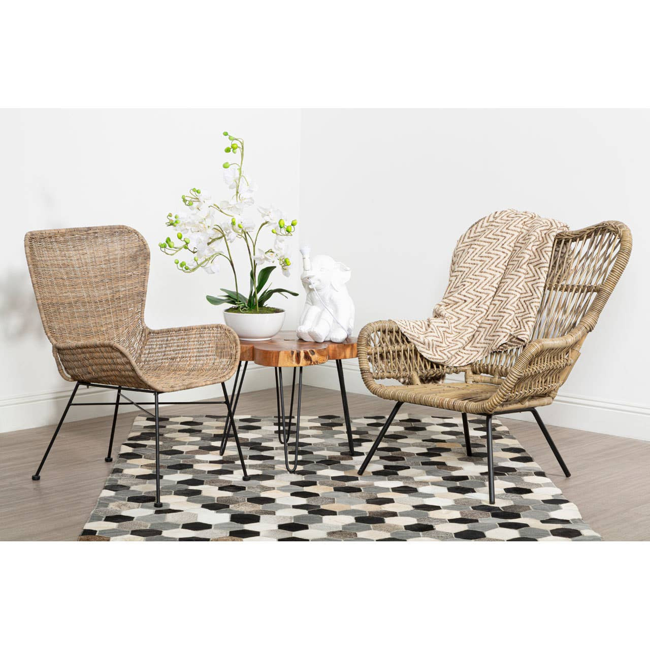 Natural rattan chair with a curved backrest and armrests, paired with a small round table, set on a patterned rug. A vase with white flowers is placed on the table, and the background features a neutral wall