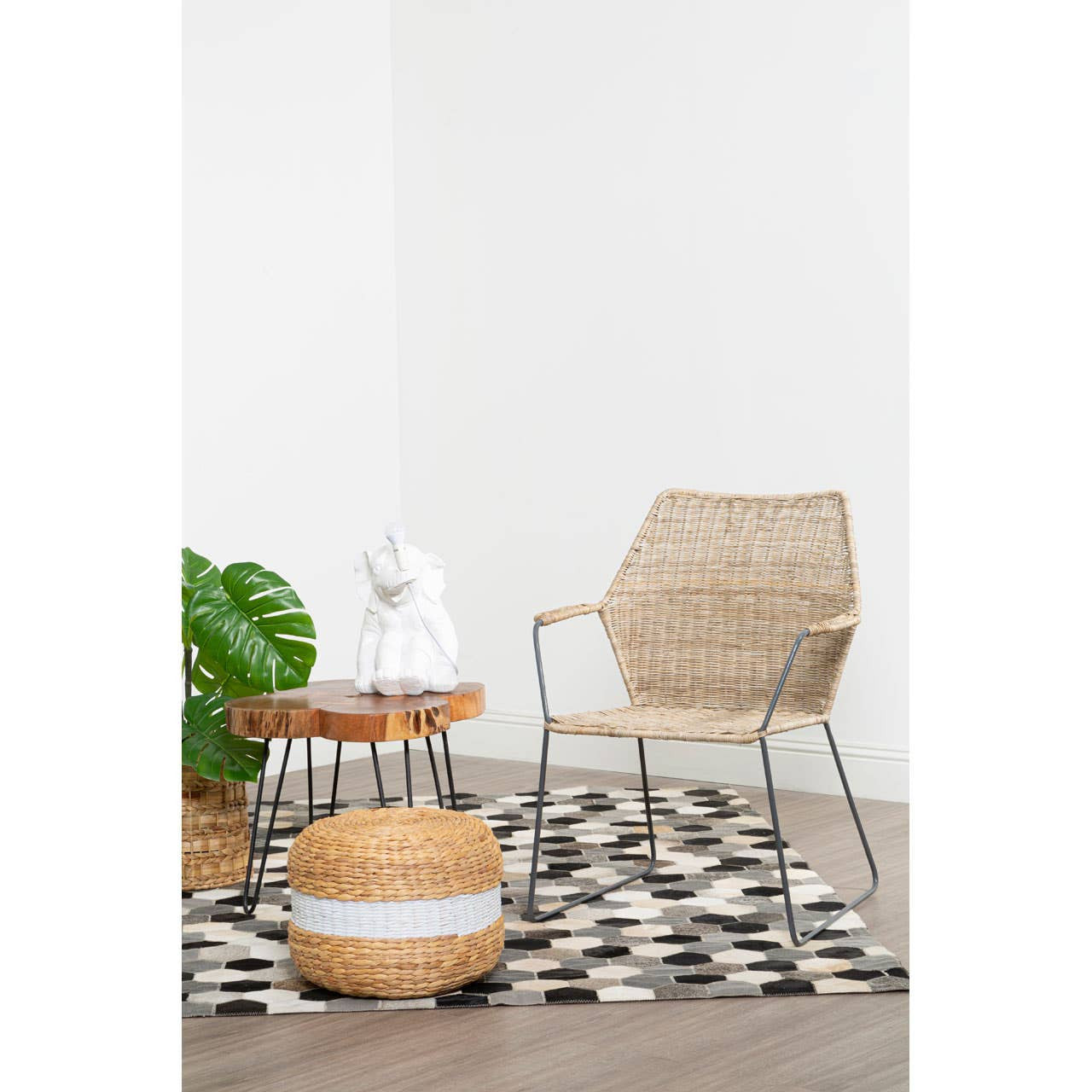 Natural rattan chair with an angled design, placed next to a round wooden side table and a patterned black and white rug. A potted green plant is visible in the background, adding a touch of greenery to the setting
