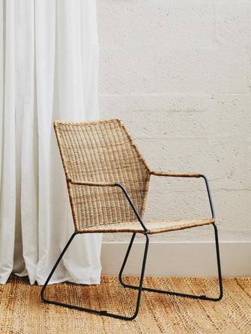 Natural rattan chair with an angled design, featuring a woven seat and backrest supported by a sleek black metal frame, placed on a wooden floor next to a white curtain