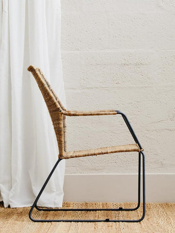 Natural rattan chair with an angled design, featuring a woven seat and backrest, supported by a sleek black metal frame. The chair is positioned beside a white curtain and on a light wooden floor, highlighting its modern aesthetic
