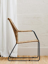 Natural rattan chair with an angled design, featuring a woven seat and backrest, supported by a sleek black metal frame. The chair is positioned beside a white curtain and on a light wooden floor, highlighting its modern aesthetic
