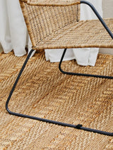 Natural rattan chair with an angled design, featuring a woven seat and backrest supported by a sleek black metal frame, placed on a textured woven rug. Light-colored curtains are visible in the background