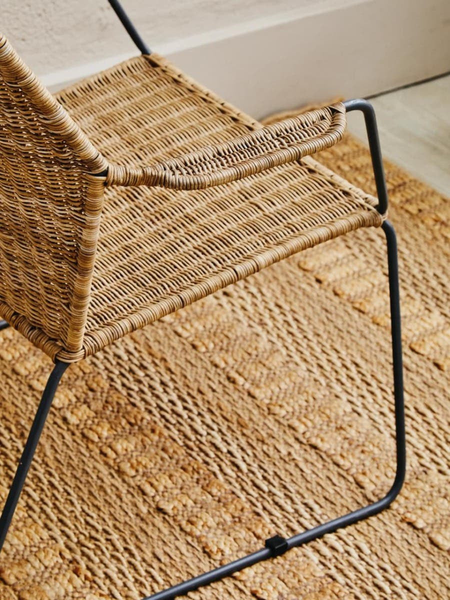 A natural rattan chair with an angled design, featuring a woven seat and backrest, supported by a sleek black metal frame. The chair is positioned on a textured rug, showcasing its modern aesthetic and natural materials