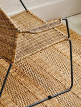 A natural rattan chair with an angled design, featuring a woven seat and backrest, supported by a sleek black metal frame. The chair is positioned on a textured rug, showcasing its modern aesthetic and natural materials