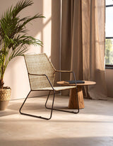 A natural rattan chair with an angled design is positioned next to a small wooden table, set against a backdrop of light-colored curtains and a potted plant. The warm sunlight casts soft shadows on the floor, enhancing the cozy atmosphere of the space