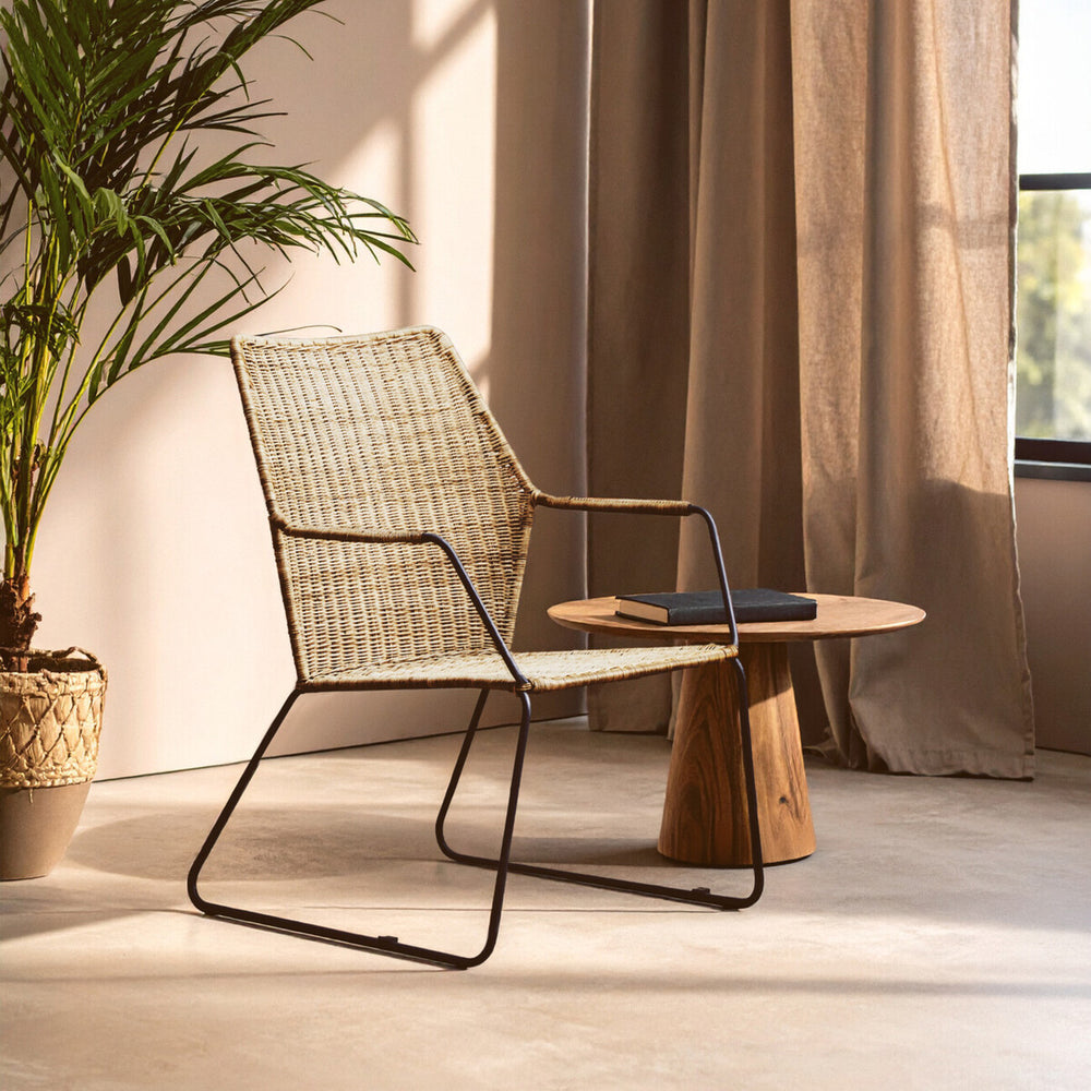 Natural rattan chair with an angled design, featuring a light-colored woven seat and backrest, supported by a sleek black metal frame. A small wooden side table is positioned next to the chair, and soft sunlight filters through sheer curtains, casting gentle shadows on the floor. A potted plant adds a touch of greenery to the scene