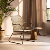 Natural rattan chair with an angled design, featuring a light-colored woven seat and backrest, supported by a sleek black metal frame. A small wooden side table is positioned next to the chair, and soft sunlight filters through sheer curtains, casting gentle shadows on the floor. A potted plant adds a touch of greenery to the scene
