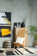 A wooden lounge chair with a light natural finish is positioned in a modern living space. The chair features a simple, slatted design and is adorned with a white cushion. Behind the chair, there is a tall wooden cabinet with a textured surface, and a large abstract painting with yellow, black, and white colors hangs on the wall. A potted plant adds a touch of greenery, and a woven