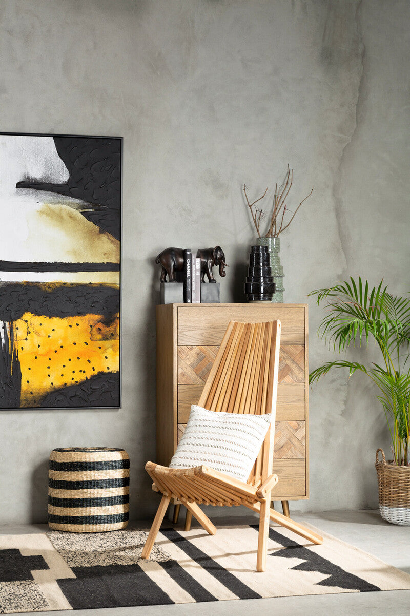 A wooden lounge chair with a light finish and a white cushion is positioned in a stylish interior. Behind it, a tall wooden cabinet displays decorative items, and a large abstract painting in black, white, and yellow hangs on the wall. A potted plant adds a touch of greenery, while a striped rug lies on the floor, enhancing the modern aesthetic of the space
