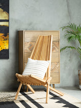 A wooden lounge chair with a natural finish, featuring a slanted backrest and a white cushion, positioned against a textured gray wall. A large piece of abstract art in warm colors is visible on the left, and a green plant adds a touch of nature to the scene