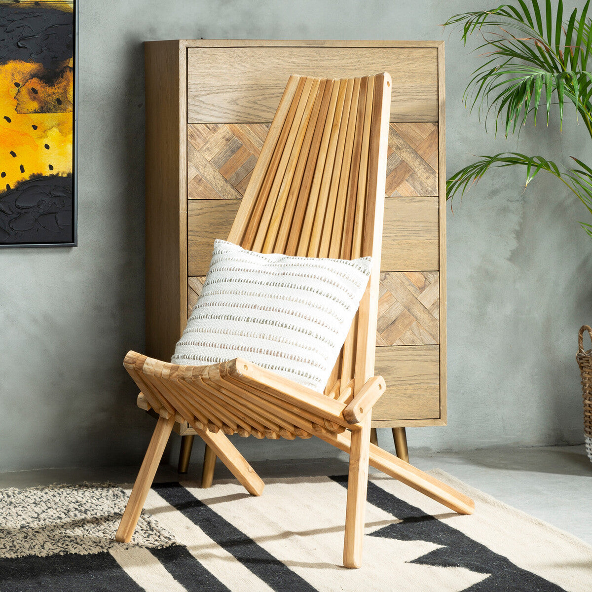 A wooden lounge chair with a slanted backrest and a light natural finish, featuring a white cushion on the seat. The chair is positioned next to a wooden cabinet against a textured wall, with a plant in the background and a patterned rug on the floor