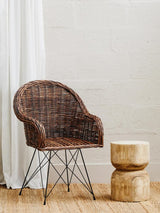 Natural croco rattan chair with a curved backrest and metal legs, positioned next to a round wooden side table, set against a light-colored wall and wooden floor