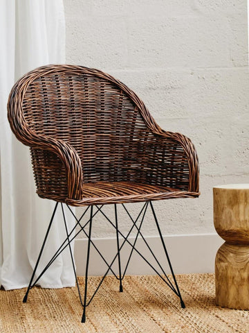 Natural rattan chair with a curved backrest and armrests, featuring a woven texture in shades of brown. The chair is supported by a metal base with a geometric design, positioned next to a round wooden side table on a light wooden floor