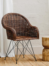 Natural rattan chair with a curved backrest and armrests, featuring a woven texture in shades of brown. The chair is supported by a metal base with a geometric design, positioned next to a round wooden side table on a light wooden floor