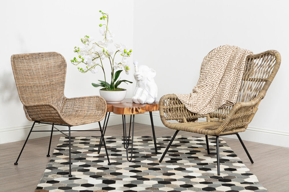 Two Manado curved design natural rattan chairs are positioned on a patterned rug, facing each other across a small round table. A white vase with green leaves sits on the table, and the background features a plain white wall. The chairs have a light brown color and a woven texture, adding a natural aesthetic to the setting