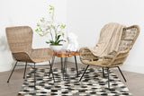 Two Manado curved design natural rattan chairs are positioned on a patterned rug, facing each other across a small round table. A white vase with green leaves sits on the table, and the background features a plain white wall. The chairs have a light brown color and a woven texture, adding a natural aesthetic to the setting