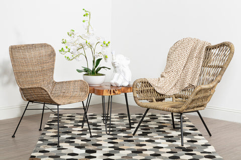 Two Manado curved design natural rattan chairs are positioned around a small round table with a white top, set on a patterned black and white rug. A vase with white flowers is placed on the table, and the background features a plain white wall. The chairs have a light brown color and a woven texture, creating a cozy and inviting atmosphere